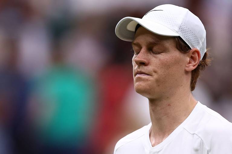 Usando boné branco e camiseta branca, o italiano Jannik Sinner, com os olhos fechados, mostra semblante de desolação nas quartas de final do Torneio de Wimbledon, na Inglaterra