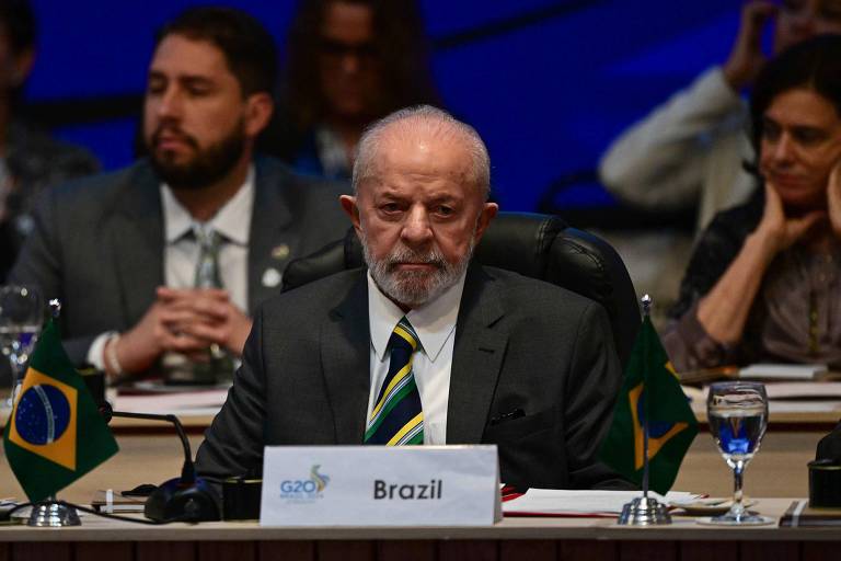 A imagem mostra um homem sentado em uma mesa durante uma reunião. Ele está usando um terno escuro e uma gravata clara. À sua frente, há uma placa com a palavra 'Brasil'. Ao fundo, são visíveis outras pessoas e bandeiras do Brasil. O ambiente parece ser uma conferência ou cúpula.