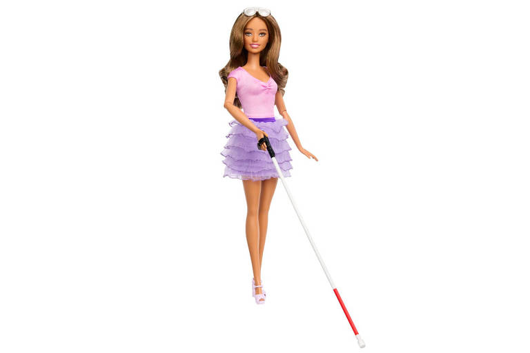 A imagem mostra uma boneca com cabelo castanho e liso, vestindo uma blusa rosa e uma saia roxa. Ela está segurando uma bengala branca com uma ponta vermelha, que é frequentemente usada por pessoas com deficiência visual.