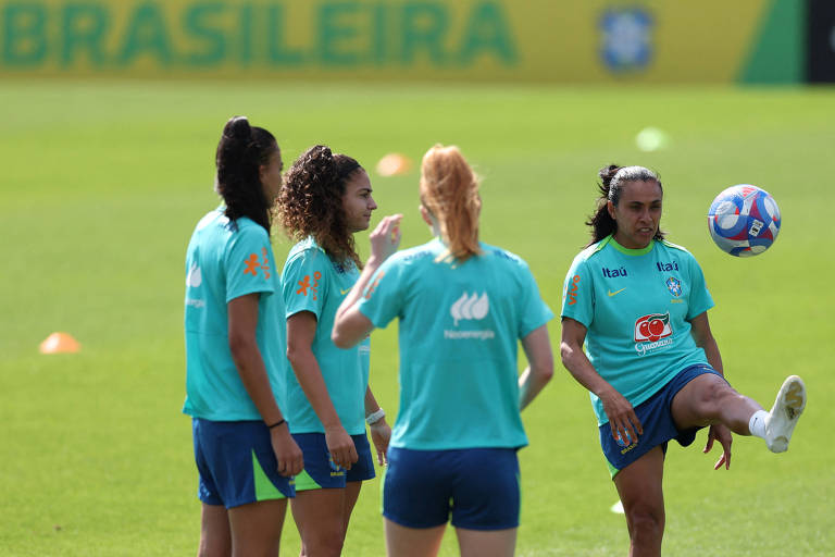 A imagem mostra quatro jogadoras da Seleção Brasileira Feminina de futebol durante um treino. Elas estão em um campo de grama, com cones de treino ao fundo. Uma jogadora está levantando a perna para chutar uma bola, enquanto as outras observam. O ambiente é ensolarado e há um banner da Confederação Brasileira de Futebol ao fundo.
