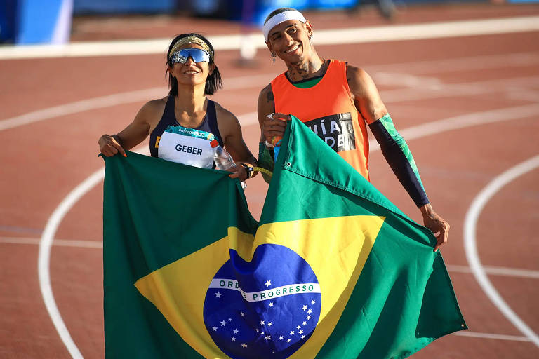 Dois atletas estão posando em uma pista de atletismo, segurando a bandeira do Brasil. Uma está vestindo uma camiseta preta e o outro uma camiseta laranja. Ambos estão sorrindo e parecem estar celebrando.
