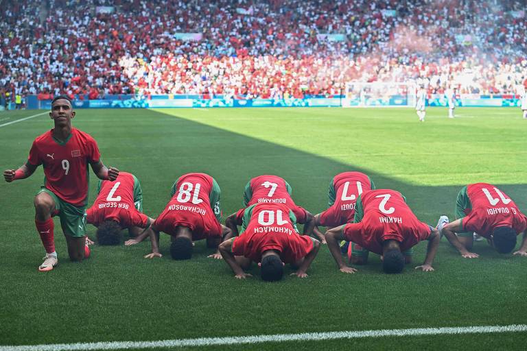 A imagem mostra jogadores da seleção de futebol do Marrocos celebrando um gol em um estádio. Um jogador, com a camisa número 9, está ajoelhado e levantando os punhos em sinal de comemoração, enquanto outros cinco jogadores, com as camisas numeradas 2, 7, 10, 17 e 18, estão se curvando em direção ao gramado, em um gesto de agradecimento. O fundo é preenchido por uma multidão de torcedores, predominantemente vestindo vermelho.
