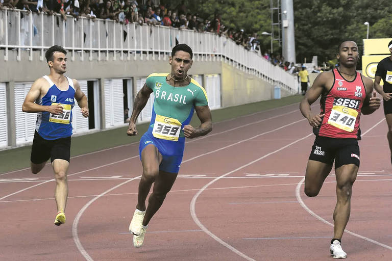 A imagem mostra três atletas competindo em uma corrida em uma pista de atletismo. O atleta à esquerda veste uma camiseta azul, o do meio está com uma camiseta verde e o da direita usa uma camiseta preta. O público pode ser visto nas arquibancadas ao fundo.
