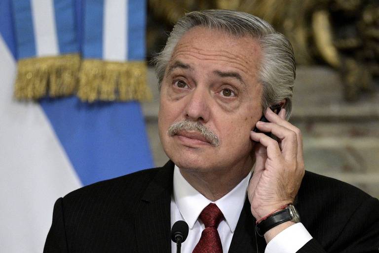 Um homem com cabelo grisalho e bigode, vestindo um terno escuro e uma gravata vermelha, está segurando um telefone próximo ao ouvido. Ao fundo, há uma bandeira da Argentina e uma decoração dourada