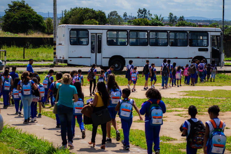 Foto mostra alunos na fila para embarcar em ônibus branco. Eles usam funiforme azul e carregam mochilas nas costas