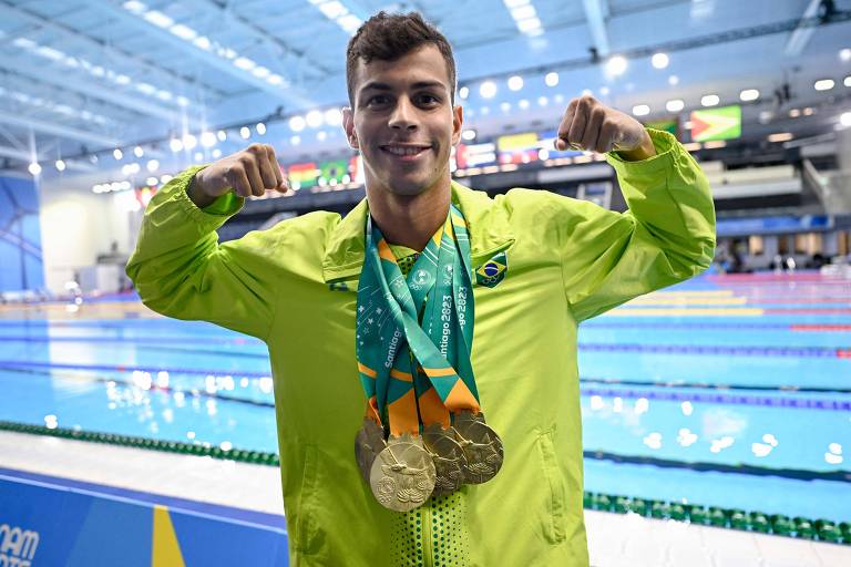 Um jovem atleta de natação está posando em frente a uma piscina. Ele usa uma jaqueta verde e exibe quatro medalhas de ouro penduradas em seu pescoço. O atleta está sorrindo e levantando os braços, mostrando os músculos.