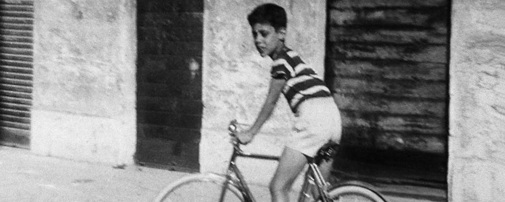 menino anda de bicicleta com camisa listrada e bermuda clara em fotografia em preto e branco
