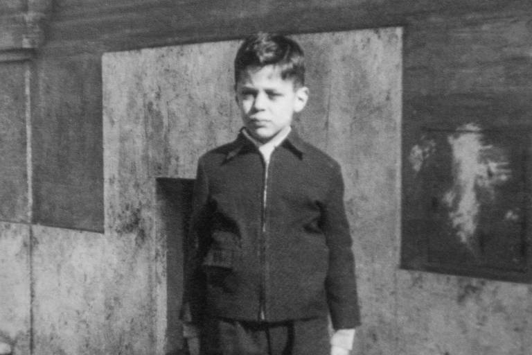 menino de blusa de manda comprida e bermuda escura, com sapatos e cabelo penteado, em fotografia em preto e branco