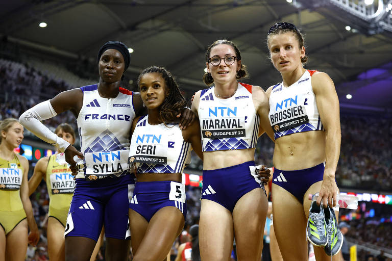 Quatro atletas femininas estão posando juntas em um ambiente de competição. Elas vestem uniformes de atletismo da França, com a bandeira francesa visível. O fundo mostra uma pista de atletismo e uma multidão. As atletas estão sorrindo e parecem estar se preparando para uma corrida.
