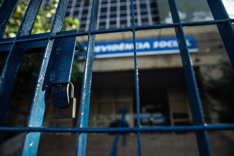 A imagem mostra um portão de metal azul com um cadeado pendurado. Ao fundo, é possível ver a fachada de um edifício, onde está escrito 'REVISÃO SOCIAL' em uma placa. O ambiente parece ser urbano, com vegetação visível ao redor do portão.
