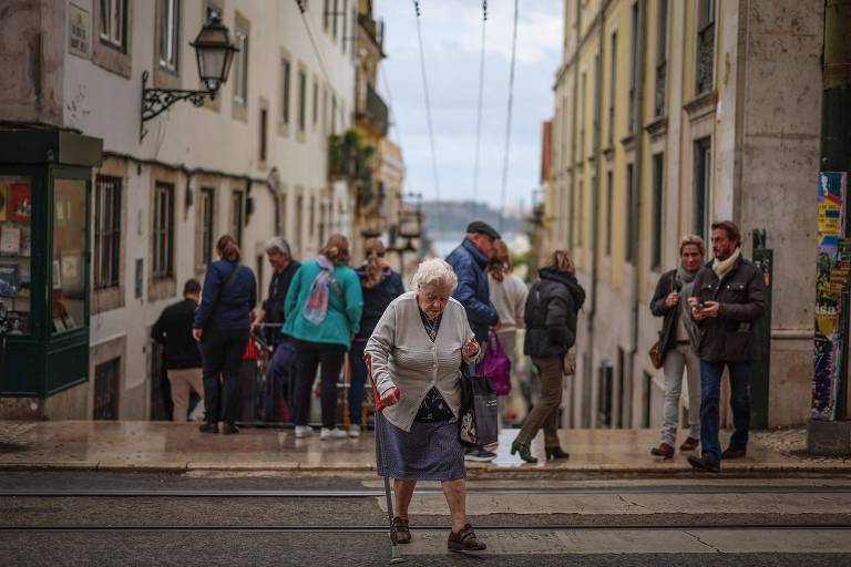 A imagem mostra uma rua em Lisboa, com várias pessoas caminhando. Em primeiro plano, uma mulher idosa com cabelo branco e usando um casaco claro caminha com a ajuda de uma bengala. Ao fundo, há um grupo de pessoas, algumas paradas e outras em movimento, em uma rua ladeada por edifícios. O céu está nublado, e há trilhos de bonde visíveis no chão.

