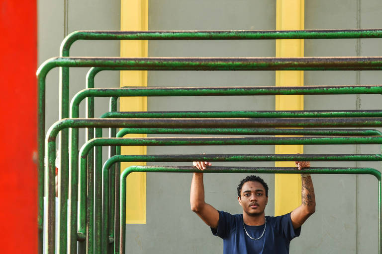 Jovem negro está segurando uma estrutura de barras verdes em uma área de recreação. Ele está posicionado em um espaço onde as barras se sobrepõem, com uma parede de fundo em cor cinza. O homem usa uma camiseta azul. 