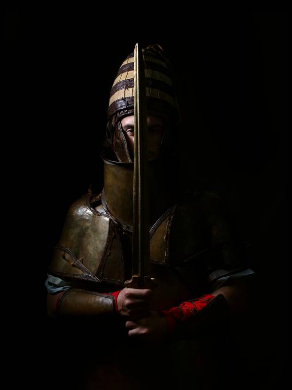 A imagem mostra um guerreiro em armadura, segurando uma espada verticalmente. O fundo é escuro, destacando a figura do guerreiro, que usa um capacete com detalhes e uma armadura metálica. As mãos do guerreiro são visíveis, uma delas com uma faixa vermelha.