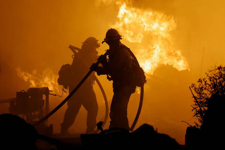 A imagem mostra bombeiros em silhueta lutando contra um incêndio. As chamas são intensas e iluminam o fundo em tons de laranja e amarelo, criando um ambiente dramático. Os bombeiros estão usando mangueiras para combater o fogo.