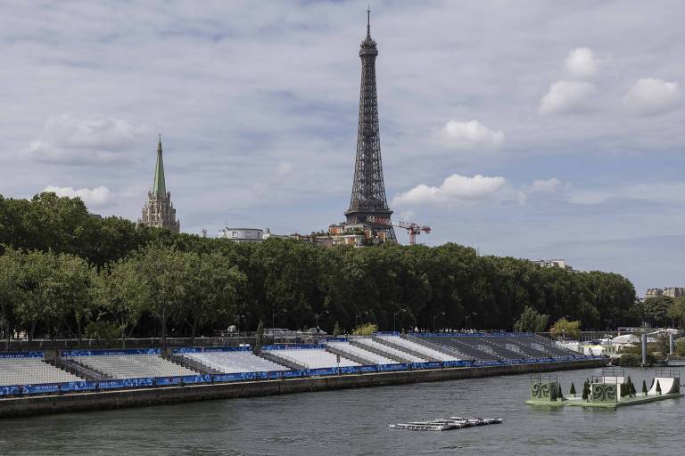 A imagem mostra a Torre Eiffel, em Paris, com um céu parcialmente nublado ao fundo. À esquerda, há uma igreja com uma torre pontuda. Na parte inferior, o rio Sena é visível, com algumas embarcações navegando. À margem do rio, há áreas verdes e estruturas de concreto.