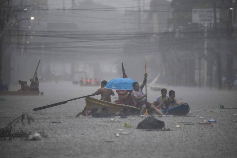 A imagem mostra várias pessoas em canoas navegando em uma rua inundada. O ambiente é sombrio, com chuva intensa e visibilidade reduzida. Algumas pessoas estão usando um guarda-chuva azul. O cenário é cercado por fios elétricos e há detritos flutuando na água.