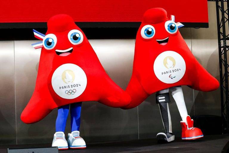 A imagem mostra dois mascotes dos Jogos Olímpicos de Paris 2024. Ambos são figuras grandes e vermelhas, com olhos grandes e expressivos. O mascote à esquerda tem um braço azul e usa um tênis branco, enquanto o da direita possui uma perna protética e um tênis vermelho. Ambos têm um emblema no peito com o texto 'PARIS 2024' e o símbolo das Olimpíadas e Paralimpíadas.
