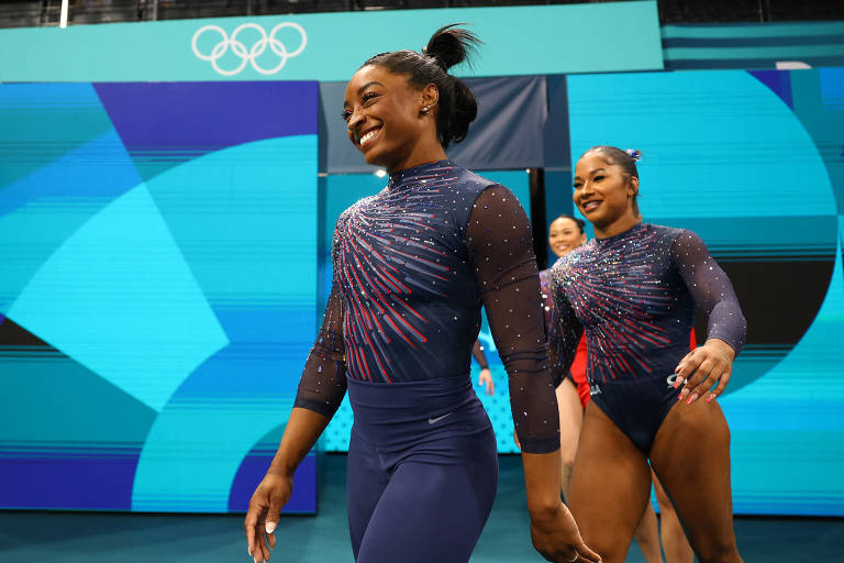 A imagem mostra duas ginastas caminhando em direção ao público durante uma competição. Ambas estão vestindo trajes de competição de mangas longas, com detalhes brilhantes. A ginasta à frente sorri, enquanto a outra também parece estar se divertindo. Ao fundo, há uma tela com o logotipo dos Jogos Olímpicos.