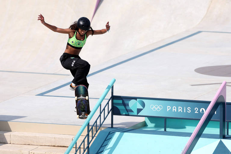 Uma skatista está realizando um truque em uma pista de skate durante os Jogos Olímpicos de Paris. Ela usa um capacete e uma blusa de alças verde, com calças pretas. A skatista está no ar, com os braços abertos, enquanto sua prancha de skate está em uma rampa. Ao fundo, há uma estrutura com a inscrição 'PARIS 2024' e o símbolo dos Jogos Olímpicos.