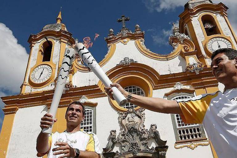 Dois homens estão segurando tochas olímpicas em frente a uma igreja histórica com uma fachada branca e detalhes em amarelo. O céu está claro e ensolarado, com algumas nuvens. A igreja possui torres com relógios e uma decoração elaborada.