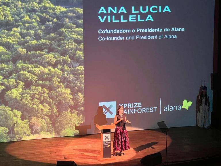 A imagem mostra Ana Lucia Villela em um palco, falando ao microfone. Ela está vestida com uma saia longa e preta com estampas coloridas. Ao fundo, há uma projeção de uma floresta verde. À direita, uma figura vestida com roupas tradicionais está em pé. O logotipo do XPRIZE Rainforest e da Alana estão visíveis na parte inferior da tela.