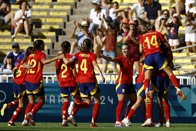 Um grupo de jogadoras da equipe feminina de futebol da Espanha celebra um gol em um estádio. As jogadoras estão vestindo uniformes vermelhos e amarelos, com algumas delas se abraçando e outras pulando de alegria. Ao fundo, há uma plateia assistindo ao jogo.
