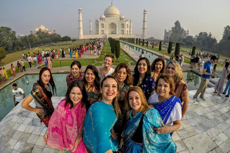 Fotografia de mulheres posando para foto em frente ao Taj Mahal, na Índia. Elas estão vestindo roupas tradicionais indianas coloridas, como saris. Ao fundo, é possível ver jardins e outros visitantes.