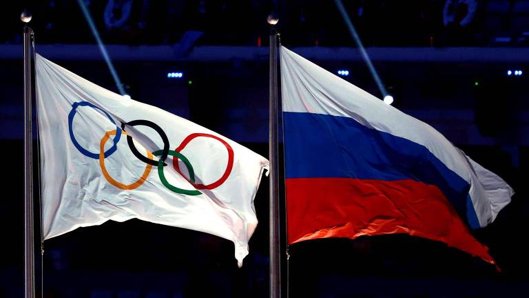 A imagem mostra duas bandeiras: à esquerda, a das Olimpíadas, uma bandeira branca com cinco círculos que se sobrepõe enfileirados nas cores azul, amarelo, preto, verde e vermelho. À direita, a bandeira da França: três faixas horizontais, uma branca, uma azul e a última de cima para baixo, vermelha.