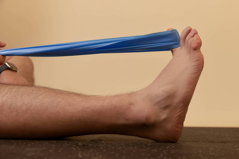 A imagem mostra uma perna de um indivíduo em posição horizontal, com um pé descalço. O pé está sendo puxado por uma faixa elástica azul, que é segurada com uma mão. O fundo é de uma cor clara, e o chão parece ser de um material escuro.
