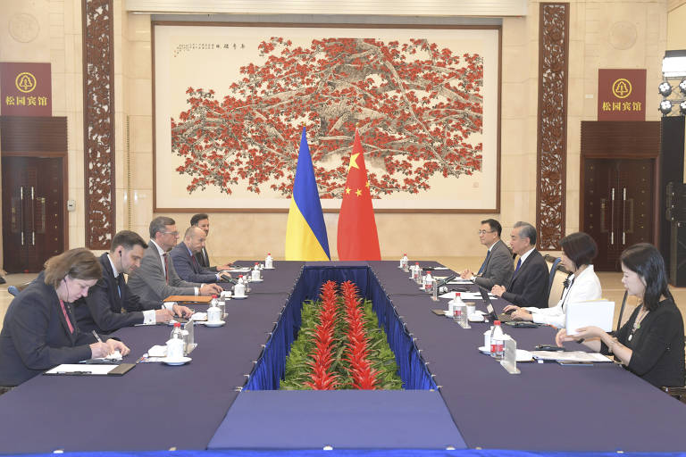 Duas delegações de quatro pessoas cada dividem uma mesa, cada uma de um lado. Ao centro, as bandeiras amarela e azul da Ucrânia e vermelha da China. Ao fundo, quadro decorativo com uma árvore e flores vermelhas.