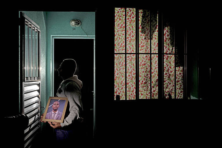 A imagem mostra uma pessoa de perfil, segurando uma moldura com uma imagem, em um ambiente escuro. À esquerda, há uma porta com uma grade e à direita, uma janela com luz filtrando através de um padrão colorido. O ambiente é predominantemente escuro, com algumas áreas iluminadas.