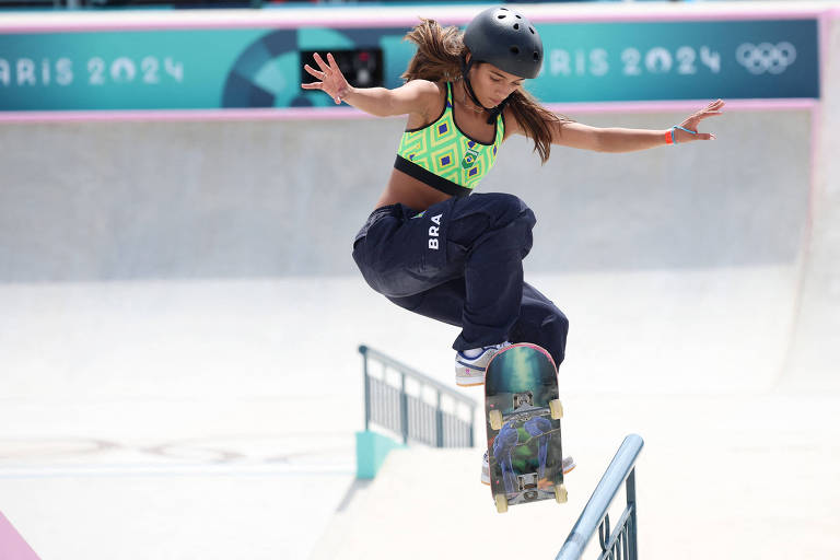 Uma skatista está realizando um salto sobre uma barra de metal em uma pista de skate. Ela usa um capacete e uma blusa verde sem alças e com estampas, combinada com calças largas. O fundo mostra uma pista de skate com elementos coloridos e a logo dos Jogos Olímpicos de Paris 2024.
