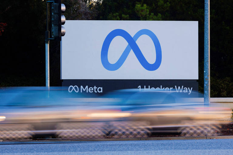 A imagem mostra uma placa da empresa Meta, com o logotipo em azul, que se assemelha a um símbolo de infinito. Abaixo do logotipo, está escrito 'Meta' e '1 Hacker Way'. Ao fundo, há um semáforo e um carro em movimento, sugerindo que a placa está localizada em uma estrada.