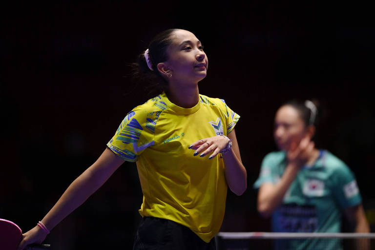 Usando camiseta amarela, a mesa-tenista Giulia Takahashi anda para a direita, com a rival ao fundo, durante competição em Busan, na Coreia do Sul
