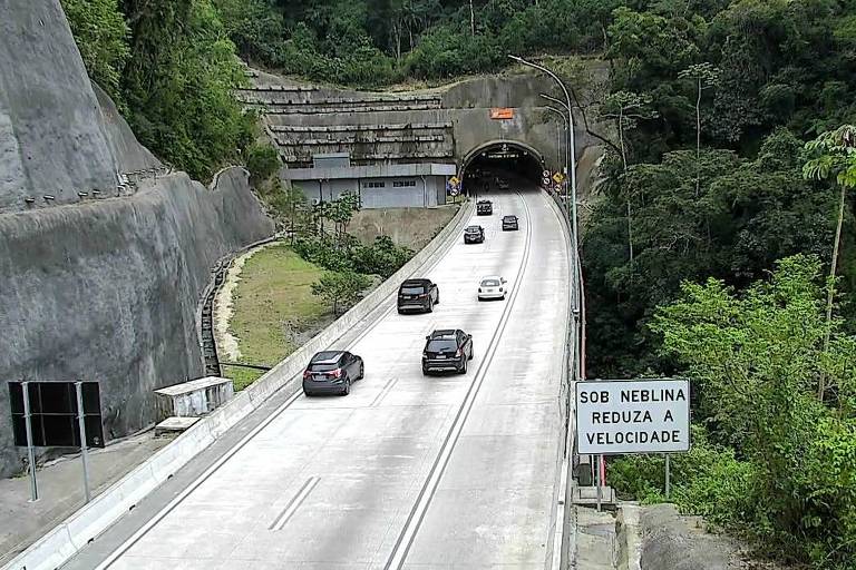 Estrada de asfalto com várias faixas de tráfego, onde alguns carros estão se aproximando de um túnel. À direita, há uma placa que indica informações sobre a rodovia
