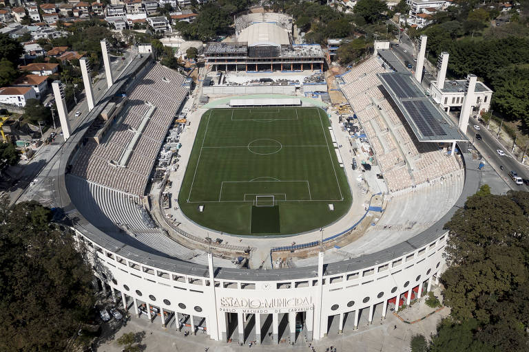 Imagem aérea do Estádio do Pacaembu, localizado em São Paulo. O estádio possui um campo de futebol visível no centro, cercado por arquibancadas. Ao fundo, há uma vista da cidade com prédios e áreas residenciais. O céu está claro e a vegetação ao redor é densa.