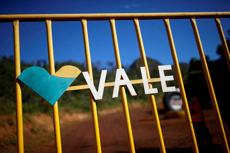 A imagem mostra um portão amarelo com a palavra 'VALE' em letras brancas. À esquerda da palavra, há a logo da empresa em azul e amarelo. O fundo é de céu azul e uma estrada de terra.