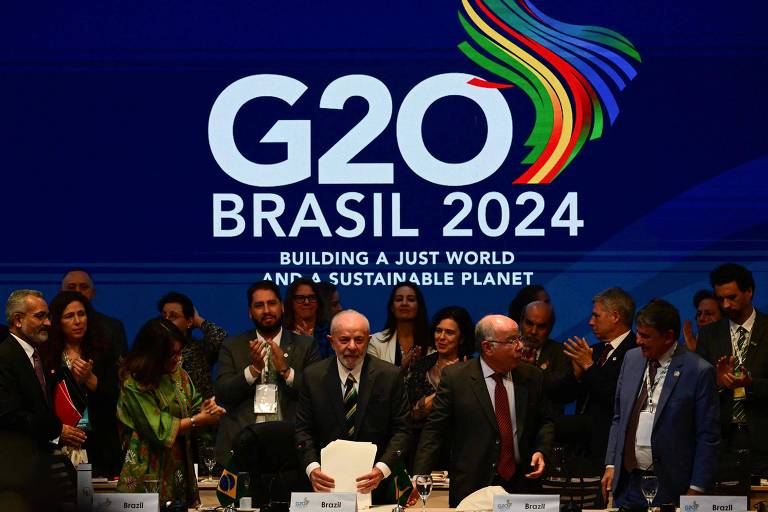 A imagem mostra uma reunião do G20, com várias pessoas em pé e sentadas ao redor de uma mesa. Ao fundo, há um grande painel com o texto 'G20 BRASIL 2024' e a frase 'BUILDING A JUST WORLD AND A SUSTAINABLE PLANET'. As pessoas parecem estar interagindo, algumas aplaudindo.