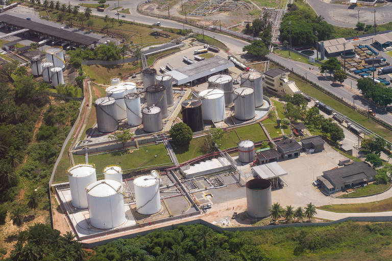 Imagem aérea de uma refinaria, com uma área industrial com tanques de armazenamento. Vários tanques brancos estão dispostos em um terreno, cercados por estradas e vegetação. Ao fundo, há mais tanques e estruturas industriais visíveis.