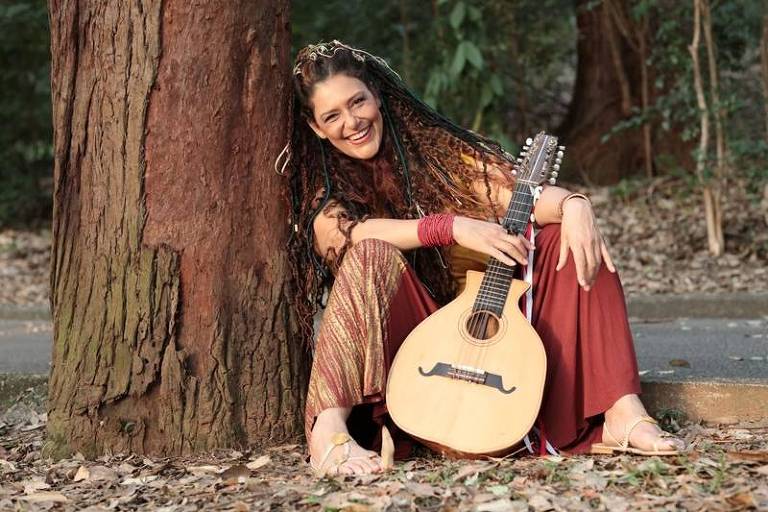 Em foto colorida, a cantora e compositora Kátya Teixeira posa para a câmera ao lado de uma árvore 