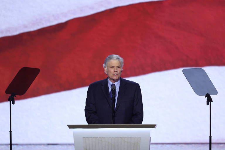 Um homem em um terno escuro fala ao microfone em um palco, com um fundo que exibe uma grande bandeira. Ele está posicionado atrás de um púlpito, com dois suportes de discurso ao seu lado.