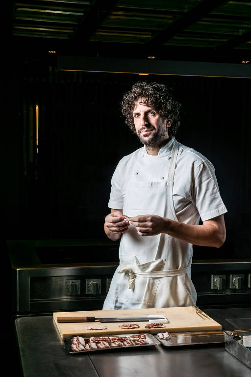 Um chef de cozinha está em pé em uma cozinha profissional, usando um uniforme branco. Ele segura um pequeno pedaço de comida com as mãos e parece estar concentrado. Ao seu redor, há uma bancada de trabalho com uma tábua de cortar e alguns ingredientes dispostos. O ambiente é escuro, com iluminação focada no chef.