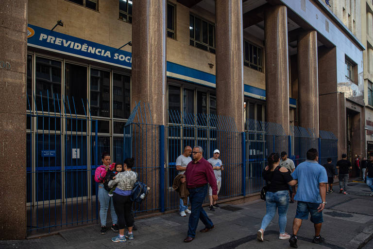 Fachada de um prédio da Previdência Social em São Paulo. Pessoas estão na calçada caminhando em frente a construção.