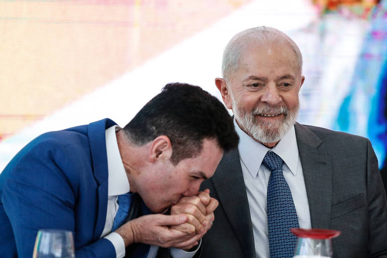 Homem de terno e gravata beija as mãos do presidente Lula, que está sorrindo, de terno e gravata 