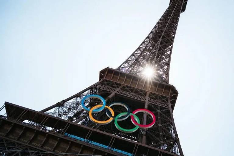 A imagem mostra a Torre Eiffel vista de baixo para cima, com o sol brilhando atrás dela. Na parte inferior da torre, estão os cinco anéis olímpicos coloridos, representando os Jogos Olímpicos.