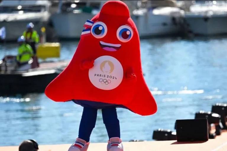 A imagem mostra a mascote das Olimpíadas de Paris 2024, que tem a forma de uma gota vermelha com um rosto sorridente. O mascote está vestido com uma camiseta branca que exibe o logotipo 'PARIS 2024'. Ao fundo, há um corpo d'água e barcos, com pessoas usando coletes de segurança visíveis.