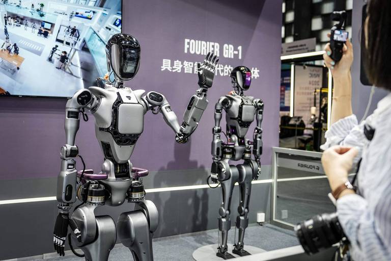 A imagem mostra dois robôs humanoides em uma exposição. Um dos robôs está acenando, enquanto o outro permanece em pé. Ao fundo, há uma tela exibindo informações sobre os robôs. Um visitante, segurando uma câmera, observa os robôs.