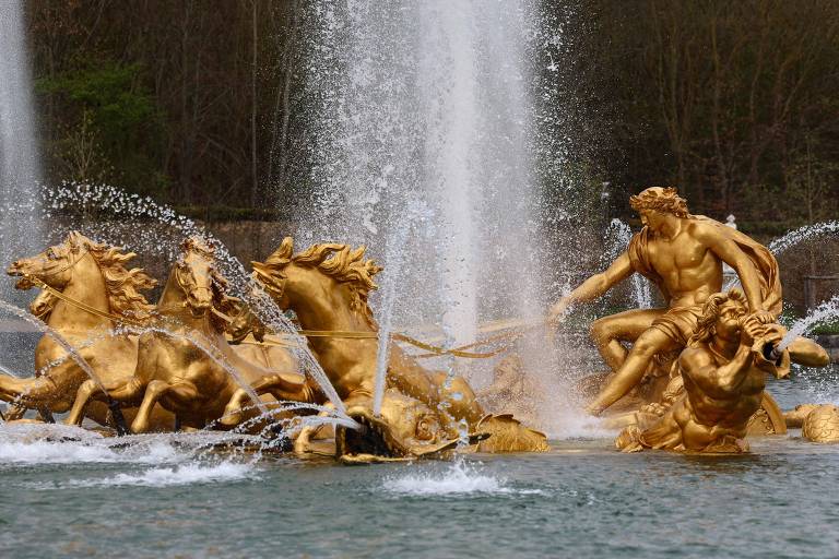 A imagem mostra uma fonte ornamentada com estátuas douradas de cavalos e figuras humanas. A água jorra em várias direções, criando um efeito visual impressionante. As estátuas estão em uma base de rochas, e o fundo é composto por árvores, sugerindo um ambiente ao ar livre.
