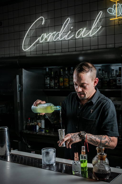 Um bartender está vertendo um líquido de uma garrafa em um copo, enquanto está em um bar. Ao fundo, há uma prateleira com várias garrafas de bebidas e uma luz neon com a palavra 'Cordial'. O bartender usa uma camisa preta e tem tatuagens visíveis nos braços.