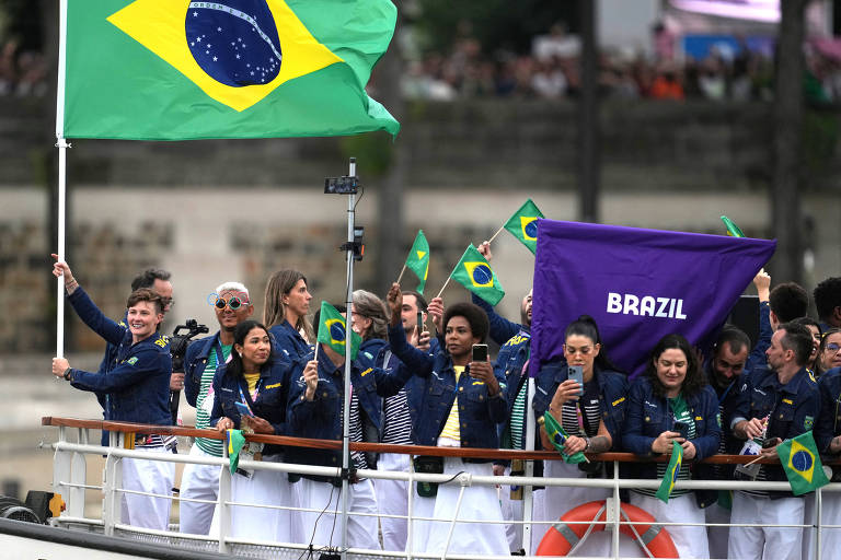 Um grupo de pessoas em um barco segurando bandeiras do Brasil e uma bandeira roxa com a palavra 'BRASIL'. As pessoas estão vestidas com jaquetas escuras e algumas estão sorrindo e acenando. Ao fundo, há uma multidão assistindo.